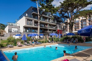 Ein Poolbereich mit Liegestühlen und Sonnenschirmen, umgeben von einem mehrstöckigen Gebäude mit Balkonen.