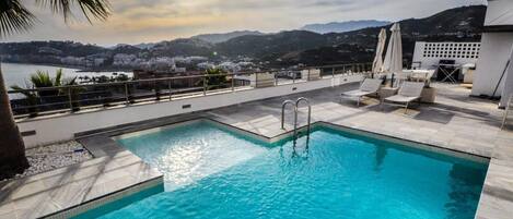 A rooftop pool with clear blue water, surrounded by lounge chairs and a view of a coastal town and mountains.