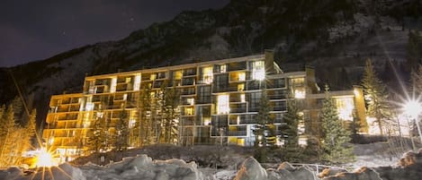 A multi-story hotel with a modern architectural design, featuring balconies and large windows, situated in a mountainous area with snow-covered terrain.