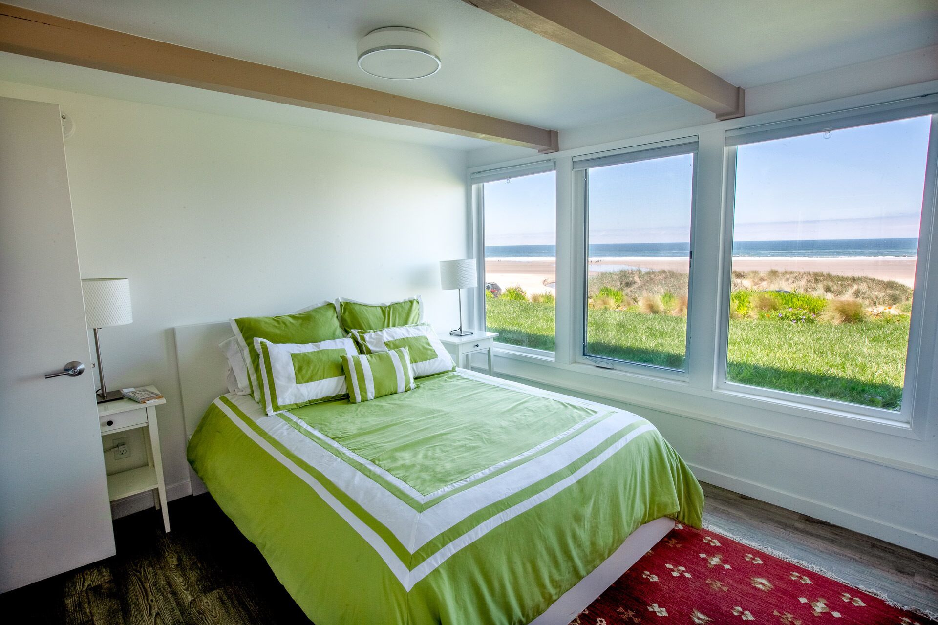 A bedroom with a large bed, a nightstand, a lamp, and a view of the beach.