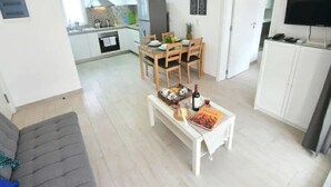 Ein modernes Wohnzimmer mit einer Couch, einem Essbereich mit Tisch für zwei Personen, einer Küche mit weißen Schränken und einem Fernseher auf einem Schrank.