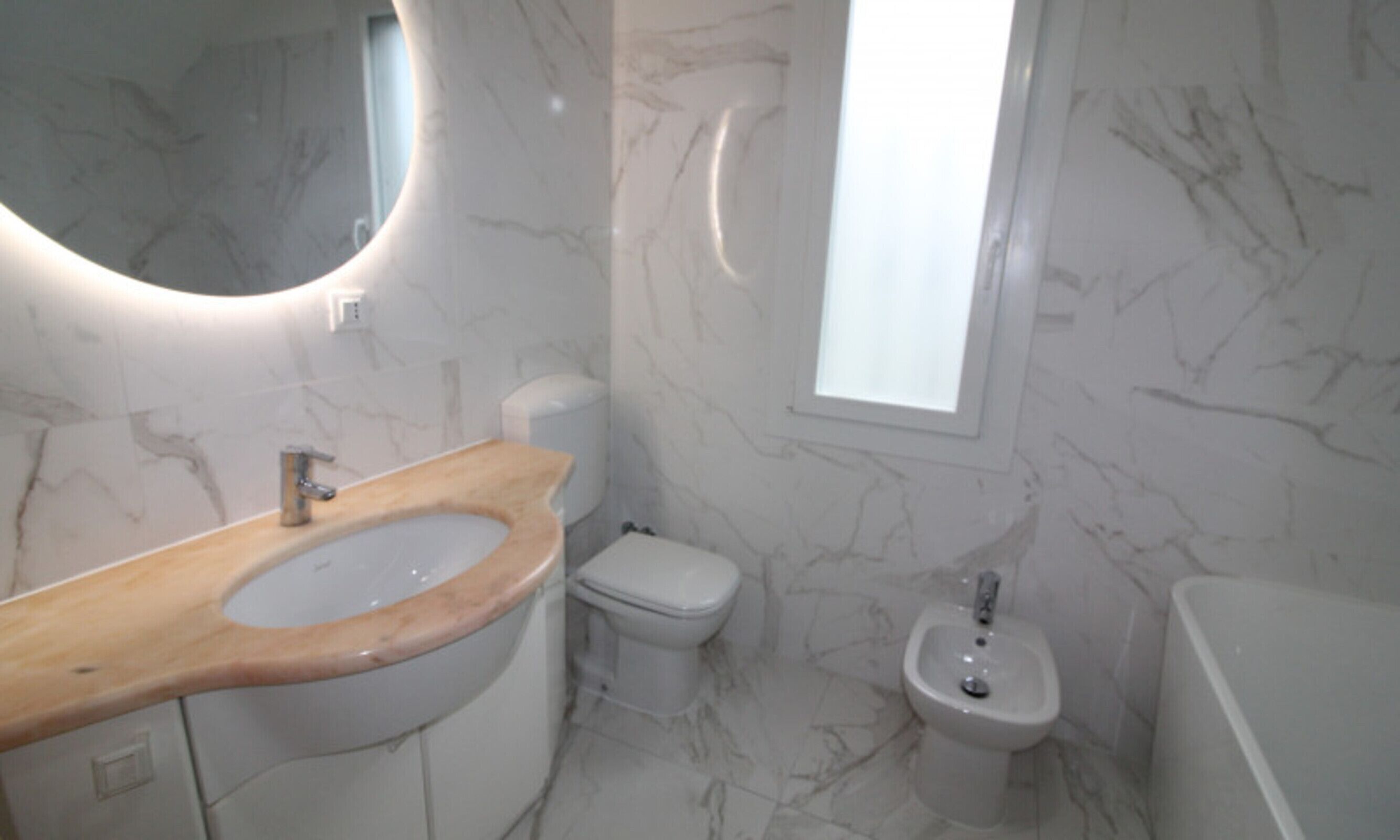 A bathroom with a marble floor, a wooden vanity, a toilet, a bidet, and a window.