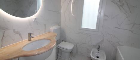 A bathroom with a marble floor, a wooden vanity, a toilet, a bidet, and a window.