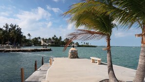 A concrete dock with benches and a palm tree, leading to a body of water with a pier and boats in the distance. - Bayside: Bayfront villa W/ free onsite boat ramp marina & trailer parking. (Islamorada)