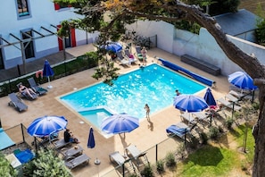 Ein Swimmingpool mit Liegestühlen und Sonnenschirmen, umgeben von einem eingezäunten Bereich, im Hintergrund ein Gebäude.