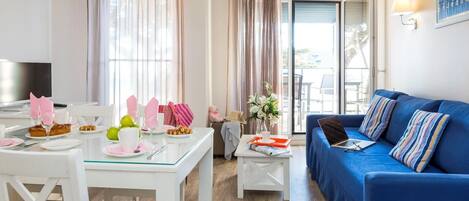 Un salon moderne avec un canapé bleu, une table à manger dressée pour deux personnes et une vue sur l’extérieur grâce à de grandes fenêtres.