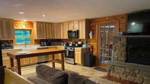 A modern kitchen with wooden cabinets, a central island, and a stone fireplace. - The Lakes Region Log Cabin (Gilford)