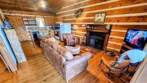 A cozy cabin interior with a wooden floor, a sofa, a fireplace, a television, and a kitchen area. - CJ's Cabin: A Secluded 1-Bedroom Retreat with Mountain Views and Hot Tub (Sieverville)