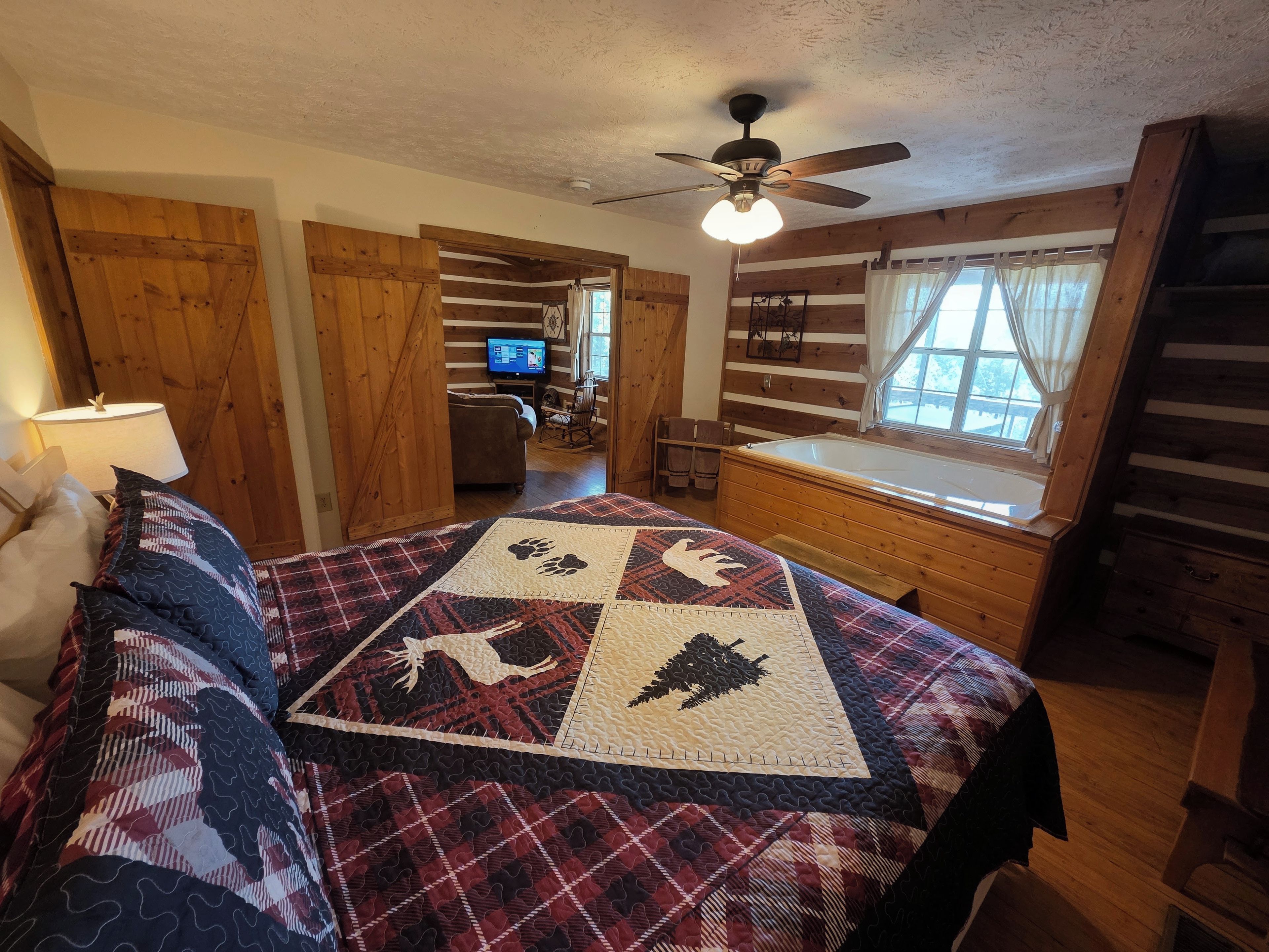 A bedroom with a bed, a ceiling fan, a window with curtains, a wooden dresser, and a television in the background.