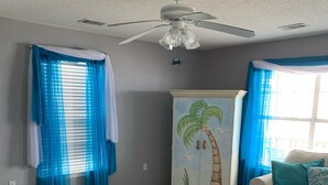 Un salón con un sofá beige, una mesa de centro de cristal, un ventilador de techo y una pared con un mural de palmeras.
