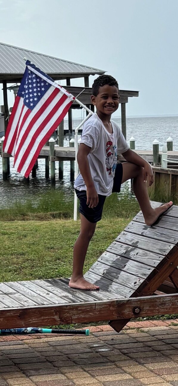 A person standing on a wooden deck with an American flag, a pier, and water in the background.