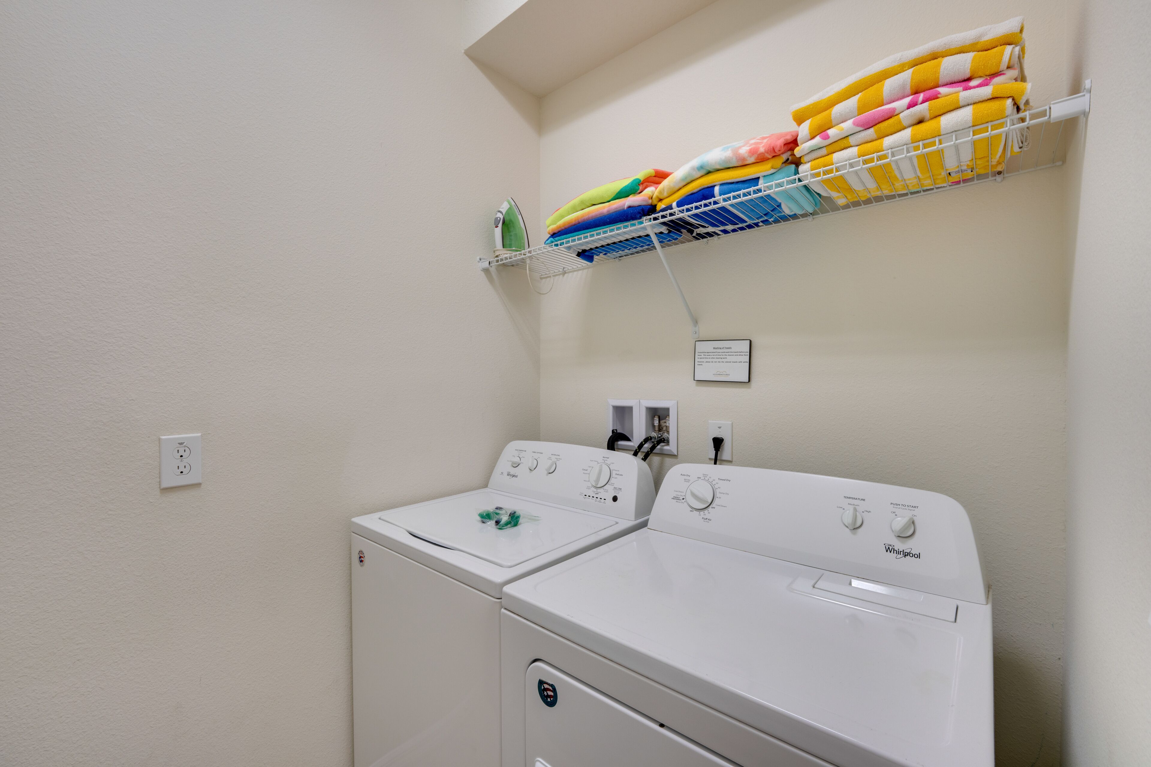 A laundry area with two white Whirlpool washers, a hanging rack with colorful towels, and a small shelf with an iron.