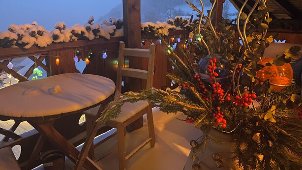 A snow-covered deck with a table and chairs, decorated with Christmas lights and greenery.