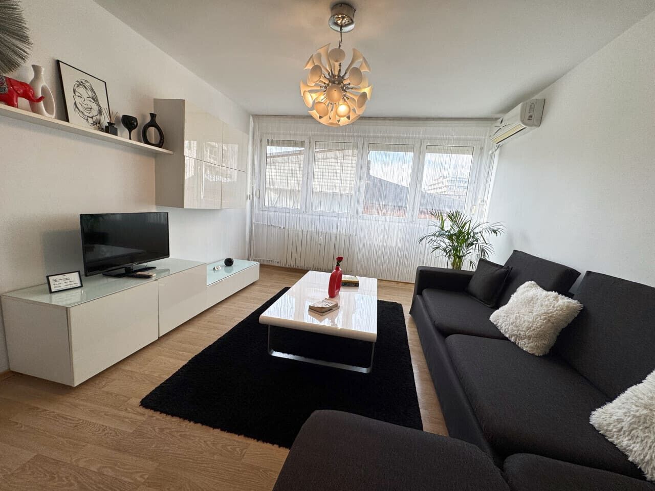 Ein modernes Wohnzimmer mit einem Flachbildfernseher, einem weißen Couchtisch, einem schwarzen Sofa und einem großen Fenster mit transparenten Vorhängen.