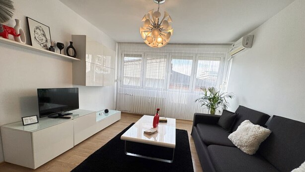 Un salón moderno con un televisor de pantalla plana, una mesa de centro blanca, un sofá negro y un ventanal con cortinas traslúcidas.