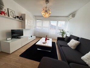 Un salon moderne doté d’un téléviseur à écran plat, d’une table basse blanche, d’un canapé noir et d’une grande fenêtre ornée de rideaux légers.