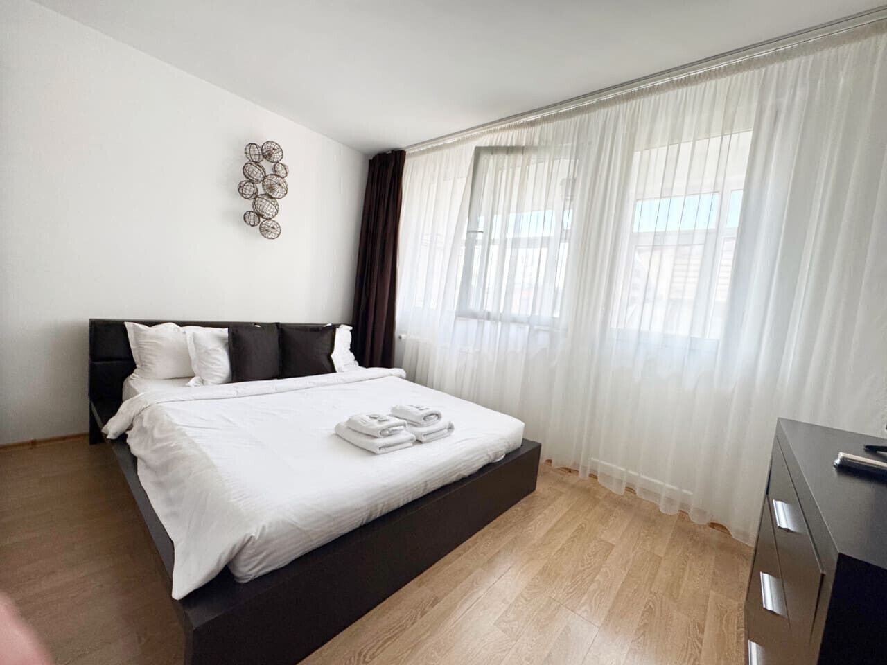 A modern bedroom with a large bed, a headboard, a bedside table, and a window with sheer curtains.