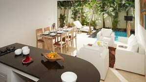 A modern kitchen with a black countertop, white bowl, and a fruit bowl. There is a dining area with a table set for four, surrounded by chairs. A white sofa and a pool are visible in the background. - 50meter from Seminyak beach,2BR Villa  (Seminyak)