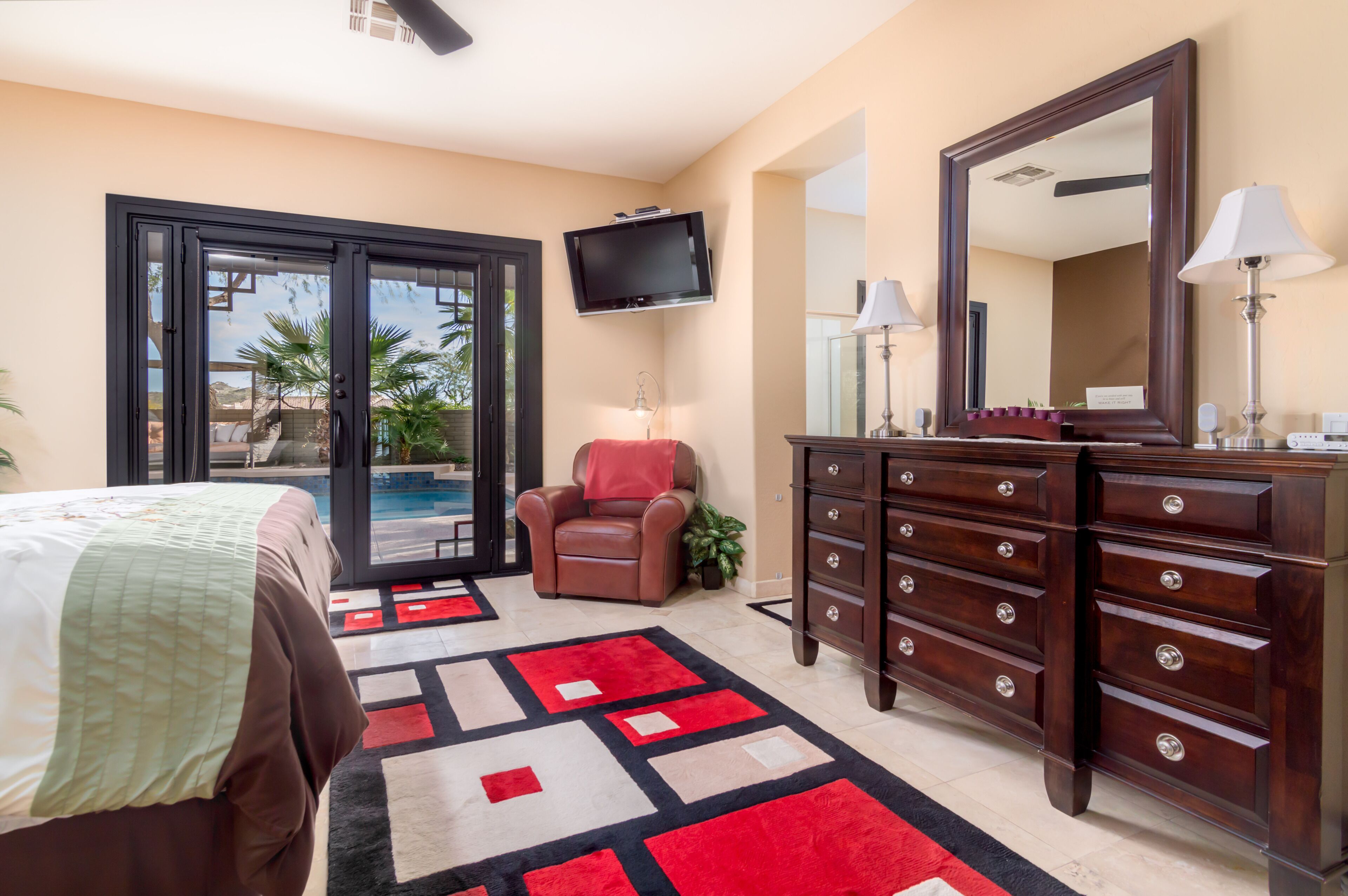 A hotel room with a bed, a desk with a lamp, a dresser, a TV mounted on the wall, and a view of the outdoors through a sliding door.