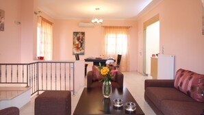A living room with a sofa, a coffee table with a vase of flowers, a dining area with a table and chairs, and a kitchen area with a refrigerator. - VILLA PANTHEON, SYVOTA BAY, LEFKADA (SYVOTA LEFKADA)