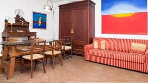 A dining area with a wooden table and chairs, a red-striped sofa, and a large painting of a sunset. - Holiday Home for 5 People, Garden, 500m till Sea, A/C, WIFI, near Forte dei Marmi (Marina Dei Ronchi)
