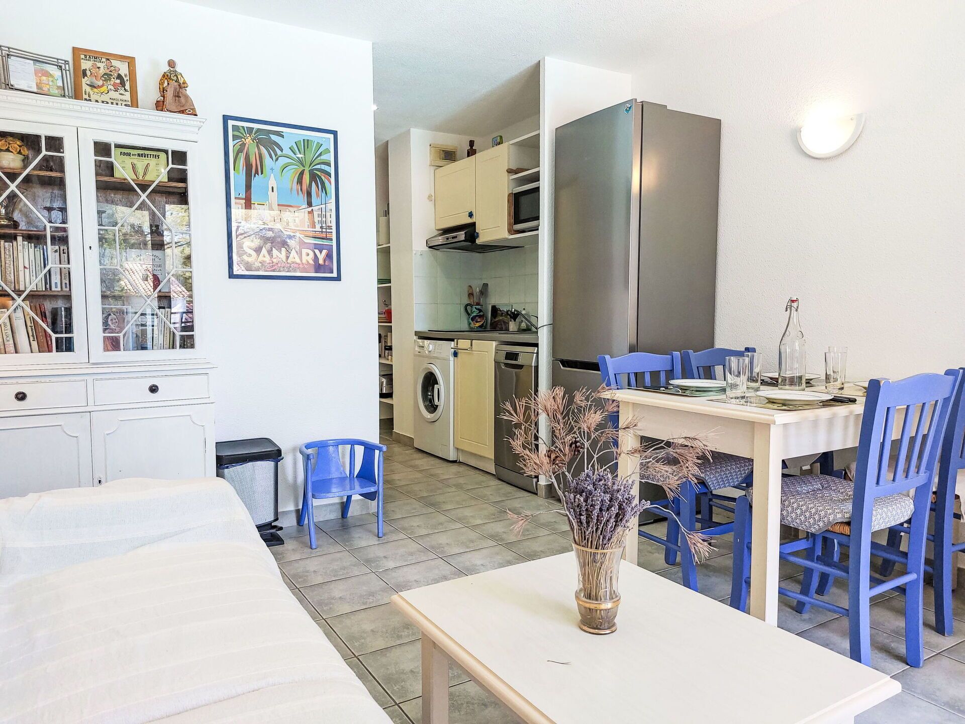 A kitchen with a stainless steel refrigerator, a dining table with blue chairs, and a vase with dried flowers.