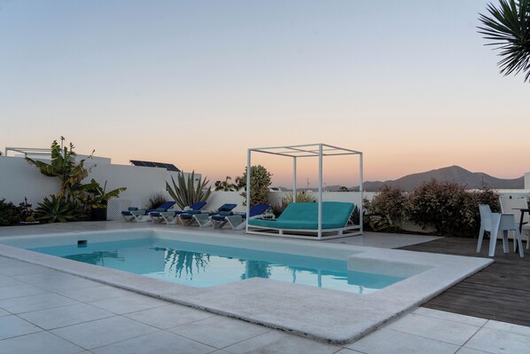 A rectangular swimming pool with a white border, surrounded by lounge chairs and a table on a wooden deck.