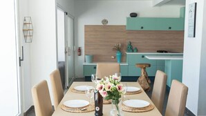 A dining area with a long table set for six, surrounded by beige chairs. A woven pendant light hangs above, and the room features a modern kitchen with teal cabinets in the background. - Villa Perla Blanca, 3 bed, free wifi, airco & heated pool (Playa Blanca)