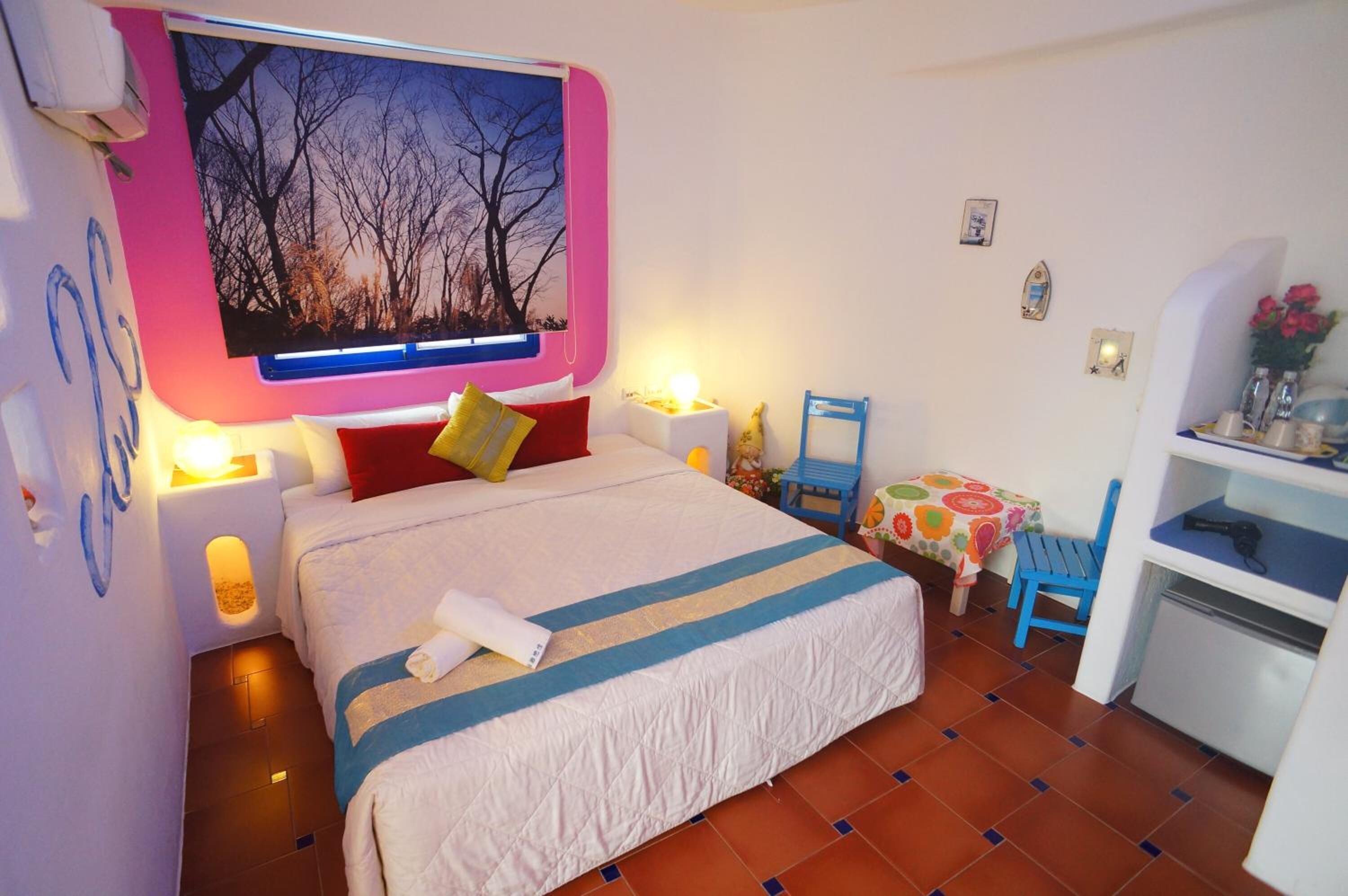 Un dormitorio con cama, mesitas de noche, una silla, una mesita pequeña y una estantería con objetos.