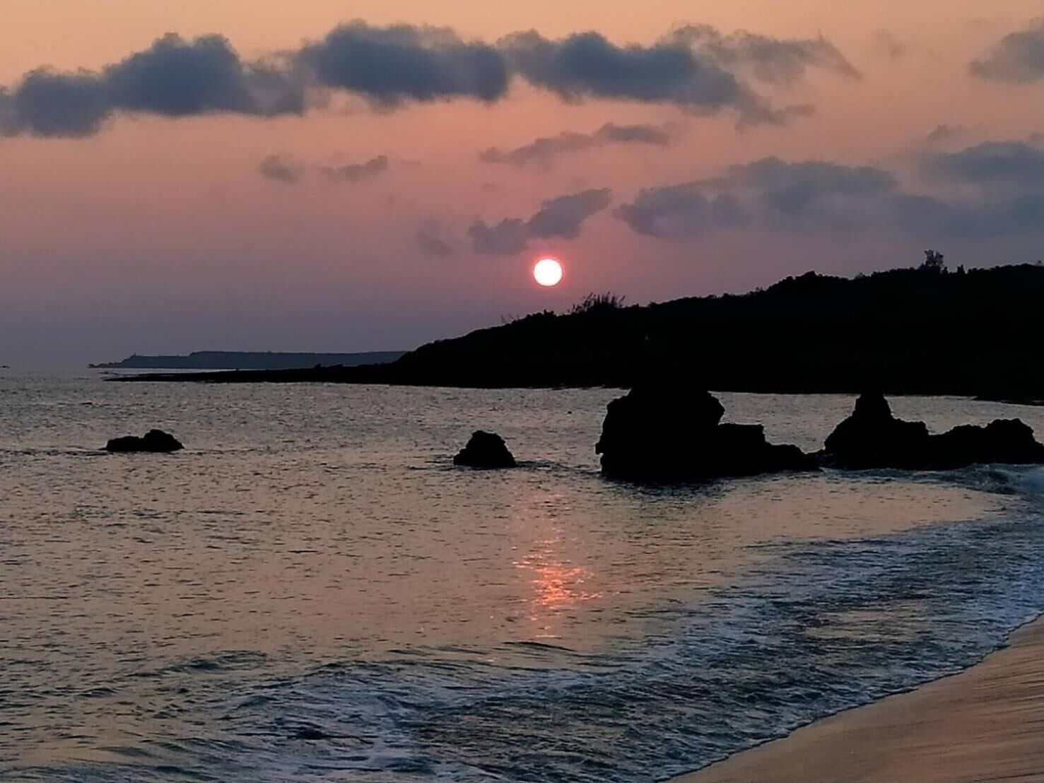 Een zonsondergang boven een rustige zee, met rotsen op de voorgrond en een verre kustlijn.