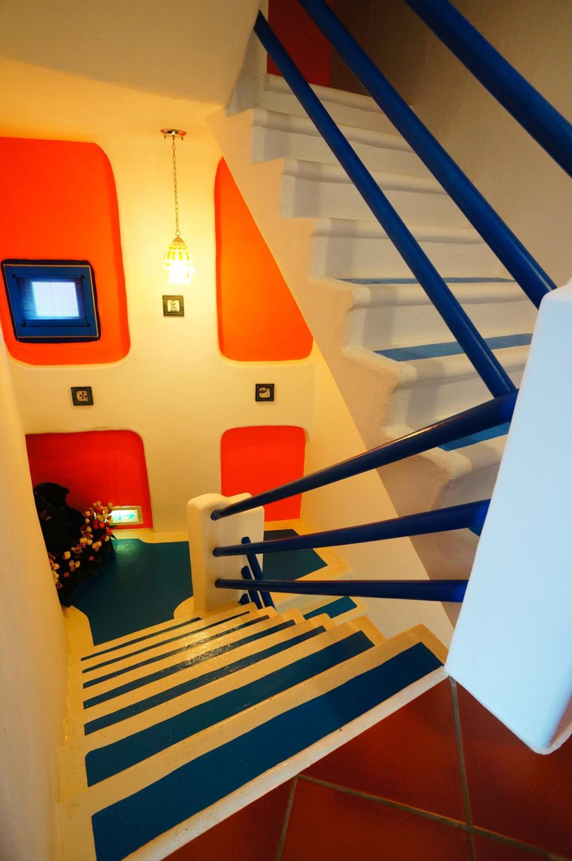Una escalera con barandas azules y peldaños blancos, que lleva a una habitación con paredes anaranjadas y un ventanal pequeño.