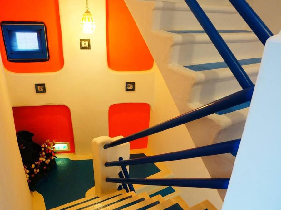 Una escalera con barandas azules y peldaños blancos, que lleva a una habitación con paredes anaranjadas y un ventanal pequeño.