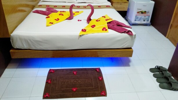 A bed with a white sheet, pink and yellow towels, and a welcome mat on the floor.