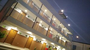 A multi-story hotel with a modern design, featuring balconies on each floor, well-lit interiors, and a series of doors likely leading to guest rooms.