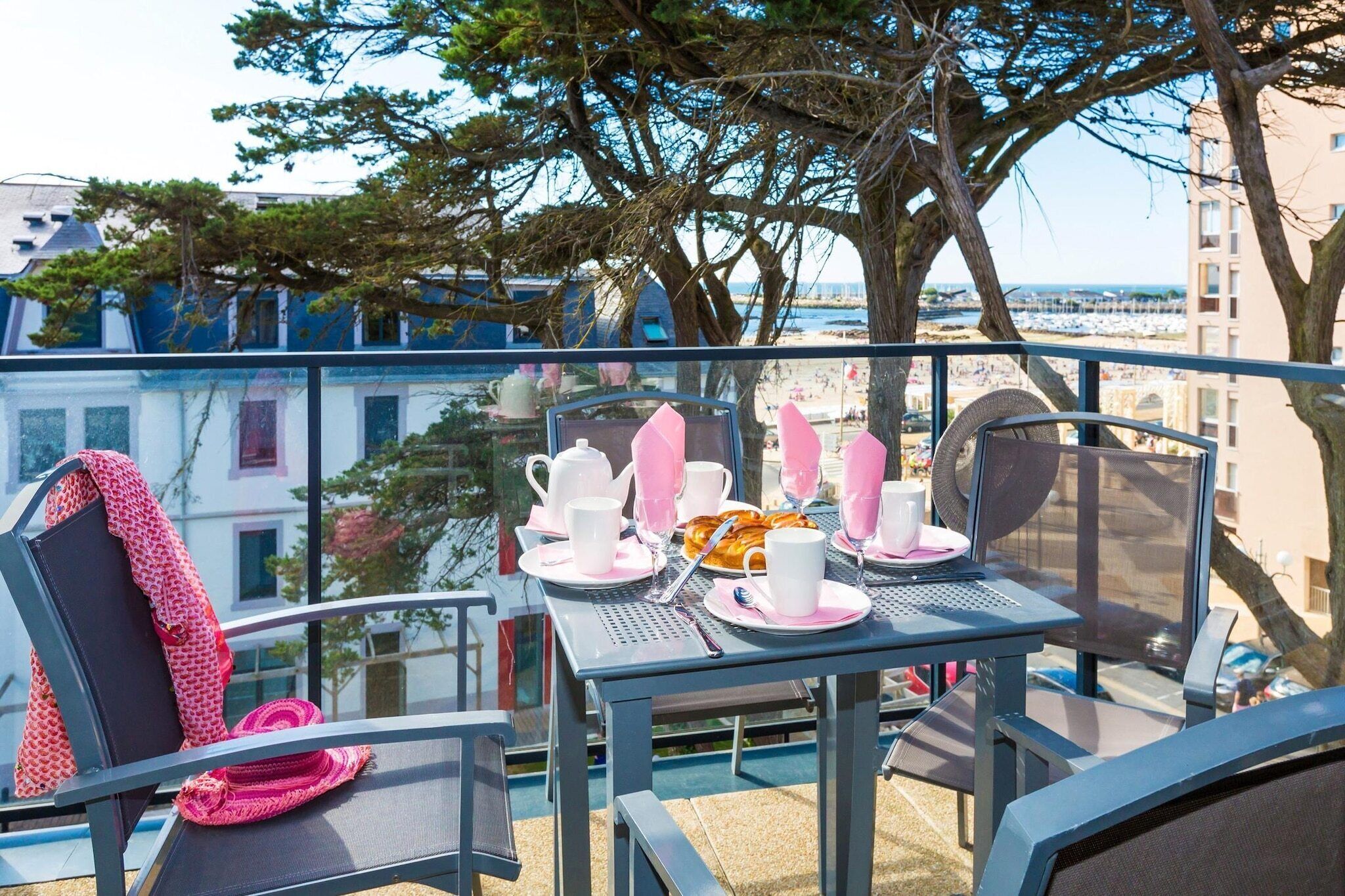 A balcony with a table set for breakfast, overlooking a beach and buildings.