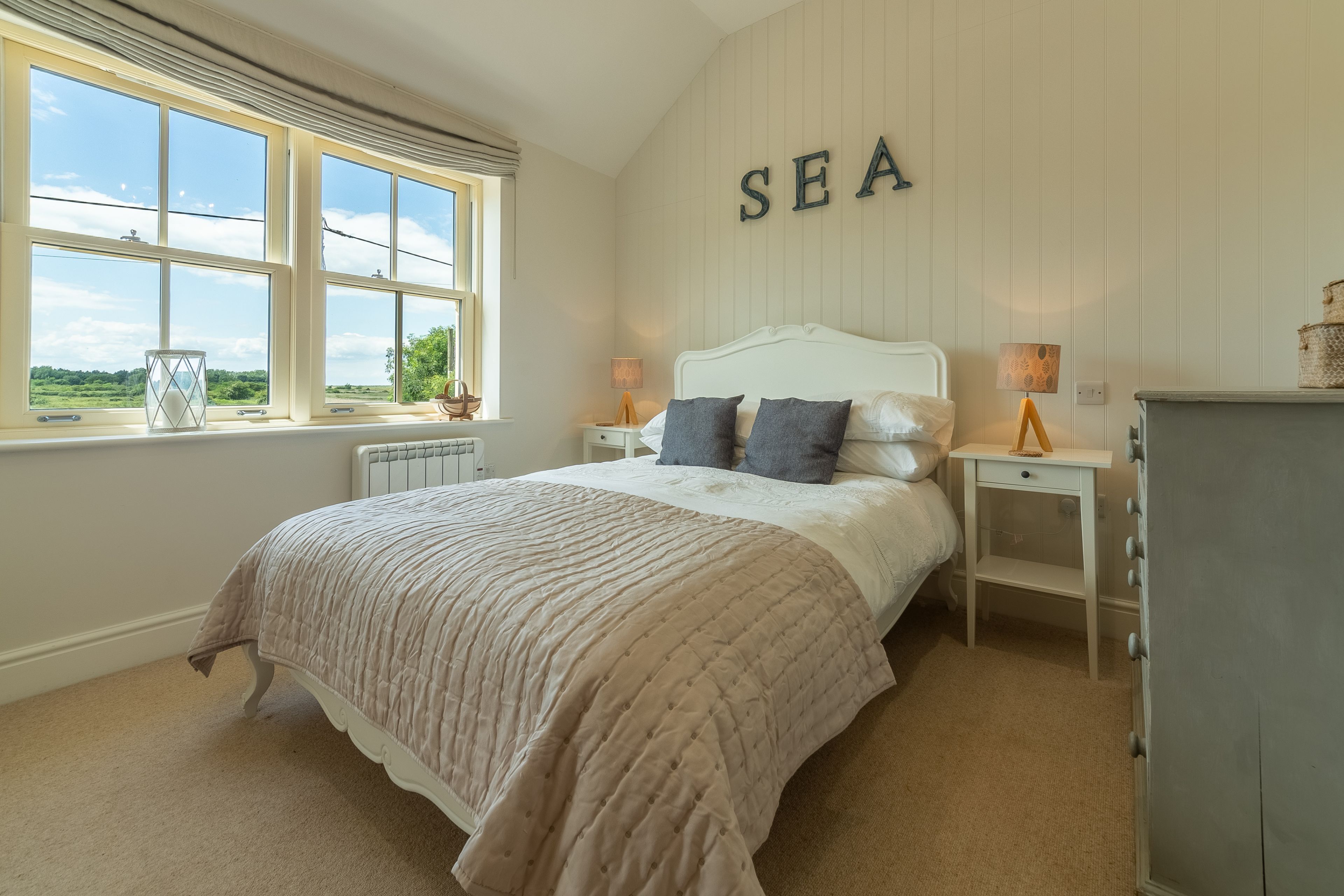 Een slaapkamer met een groot bed, een raam met uitzicht op zee, een nachtkastje met een lamp en een dressoir.