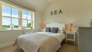 A bedroom with a large bed, a window with a view of the sea, a bedside table with a lamp, and a dresser.
