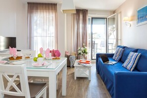 Ein modernes Wohnzimmer mit einem blauen Sofa, einem Esstisch gedeckt mit Tellern und Gläsern sowie einem Fernseher auf einem weißen Schrank.