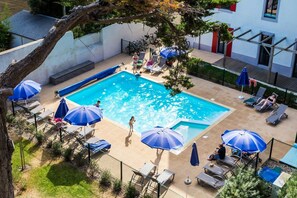 A swimming pool with sun loungers and umbrellas, surrounded by a fence and greenery.