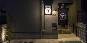 A corridor with a sign indicating a TRIPPOYOSUKE B entrance, a T-shaped logo on the wall, and a metal gate on the right. - Trip Pod Yoshizuka B (Fukuoka)