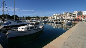 Un puerto deportivo con varias embarcaciones amarradas, un paseo marítimo pavimentado y edificios al fondo.