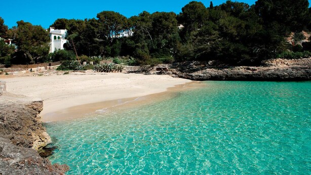 Una playa con agua turquesa cristalina, orilla arenosa y un edificio blanco con arcos al fondo.