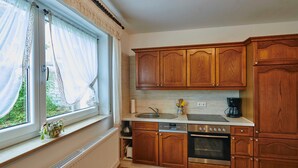 A kitchen with wooden cabinets, a built-in oven, a dishwasher, and a window with sheer curtains. - FEWO Geisbergblick, Germany (Strullendorf)