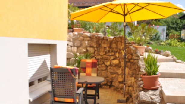 An outdoor patio with a stone wall, a yellow umbrella, and a table with chairs. - FEWO Geisbergblick, Germany (Strullendorf)