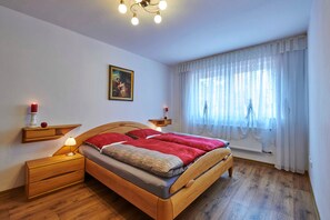 A bedroom with a wooden bed, bedside tables, a nightstand, a wall-mounted shelf with a red candle, a framed picture, and a window with sheer curtains. - FEWO Geisbergblick, Germany (Strullendorf)