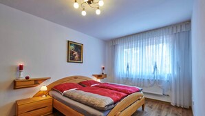 A bedroom with a wooden bed, bedside tables, a nightstand, a wall-mounted shelf with a red candle, a framed picture, and a window with sheer curtains. - FEWO Geisbergblick, Germany (Strullendorf)