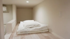 A small, empty room with a bed, a radiator, and a light switch on the wall. - Trip Pod Higashihie (Fukuoka)