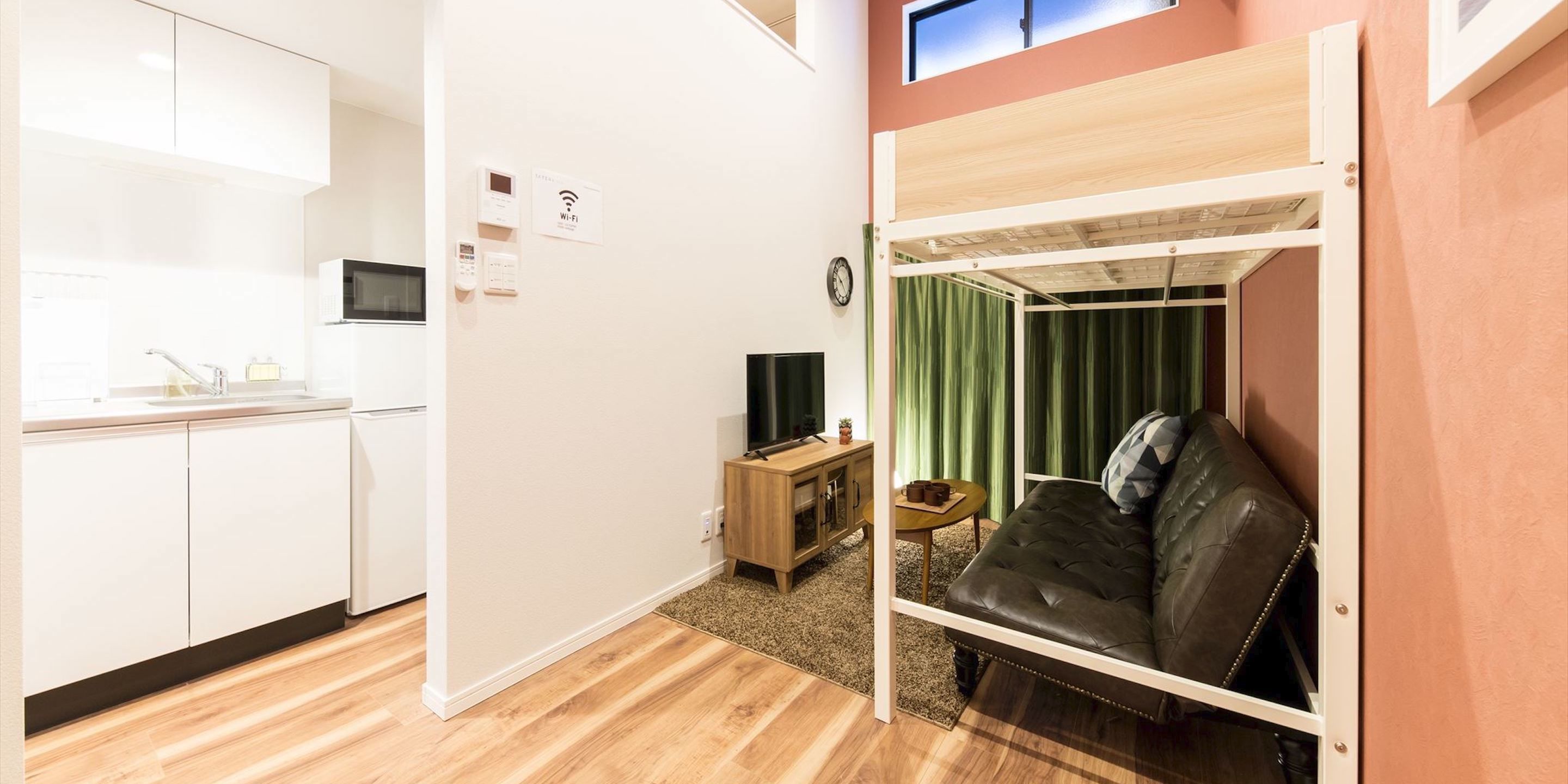 A compact room with a kitchenette, a bed with a green curtain backdrop, and a TV on a wooden side table.