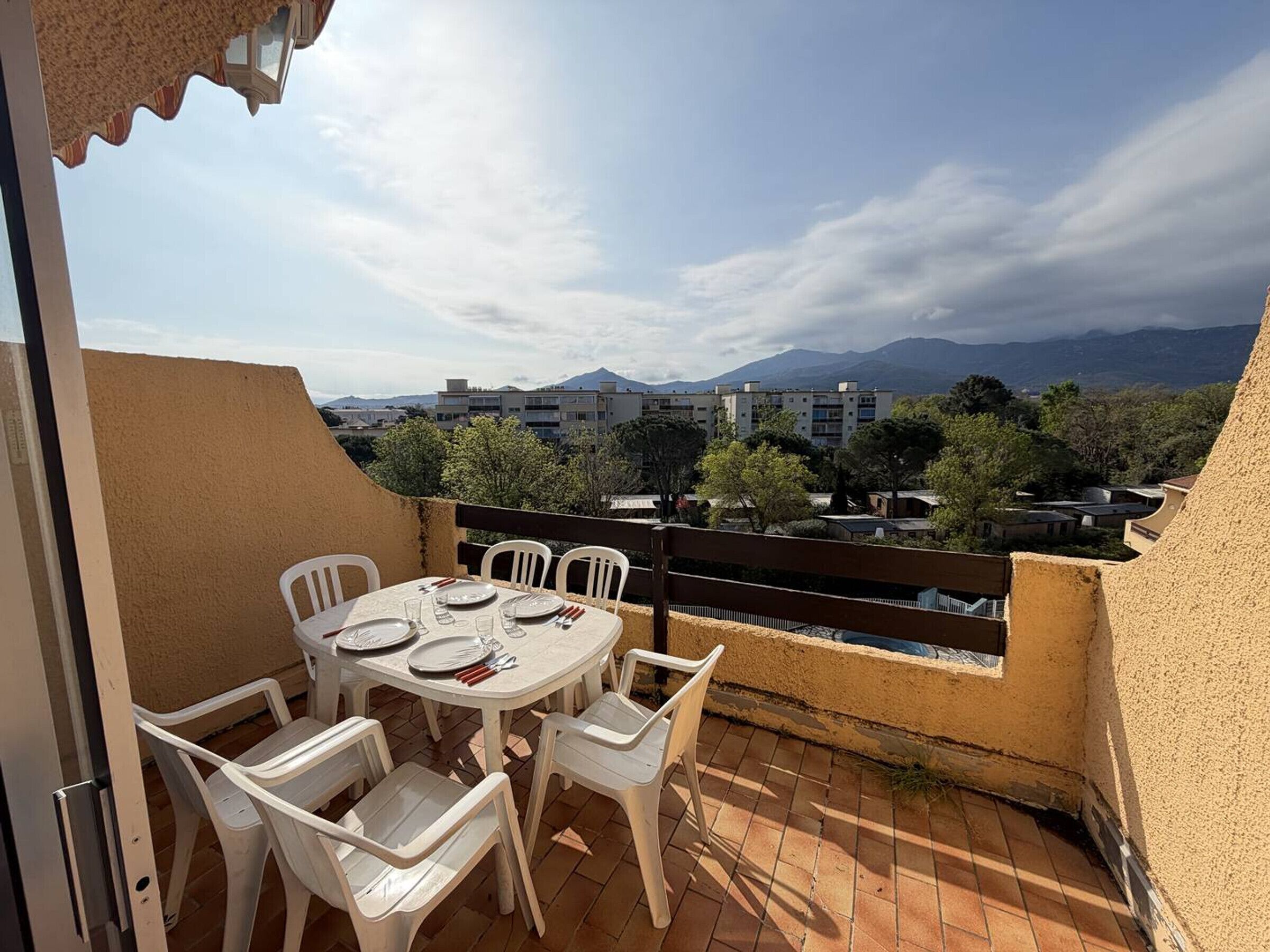 A terrace with a table set for two, white plastic chairs, and a view of buildings and mountains in the background.