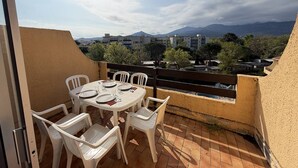 Una terraza con una mesa dispuesta para dos, sillas de plástico blancas y vistas a edificios y montañas al fondo.
